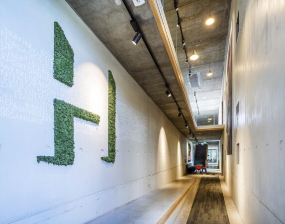 Moderne, betonnen gang in het Fred. Roeskestraat-gebouw met groene moswand in de vorm van een “H” en een trap aan het einde.